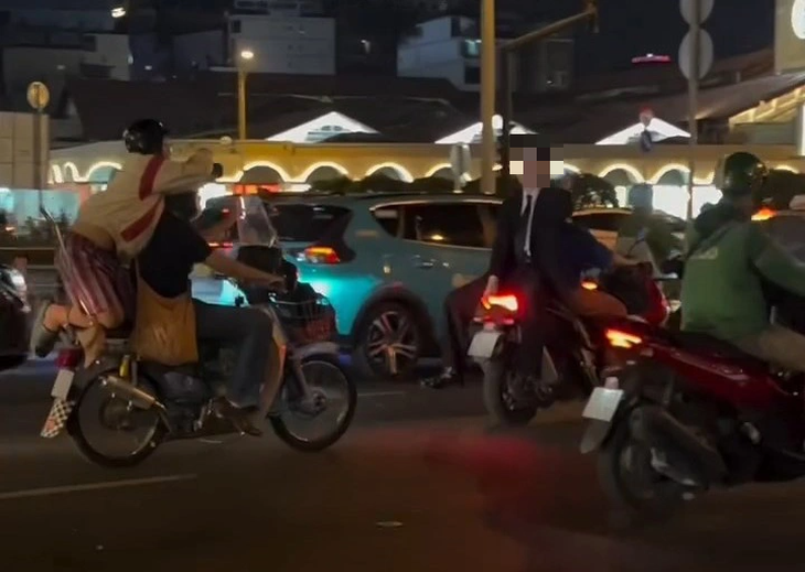 A video screenshot shows a girl kneeling on a motorbike to film a German man wearing no helmet and sitting back to back with another motorbike driver.