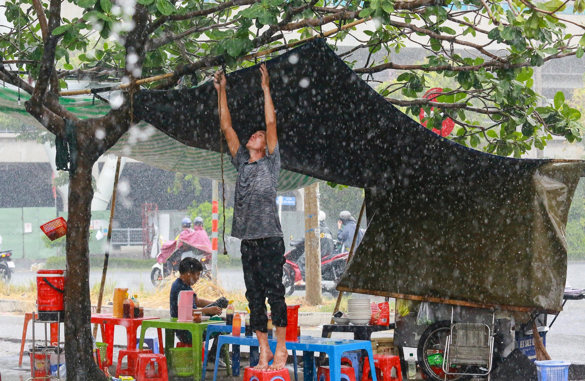A street food vendor adjusts a plastic sheet to protect his food stall from the rain. Photo: Le Phan / Tuoi Tre
