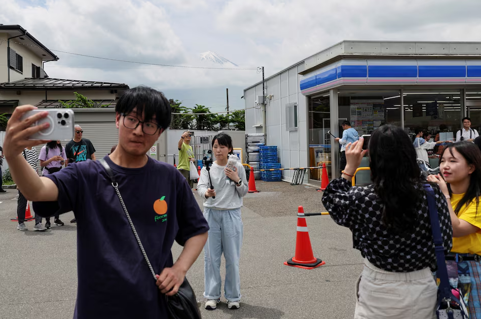 A tourist poses for a photo of Mount Fuji appearing over a convenience store after a barrier to block the popular tourist spot was installed, in Fujikawaguchiko town, Yamanashi prefecture, Japan, May 21, 2024. Photo: Reuters