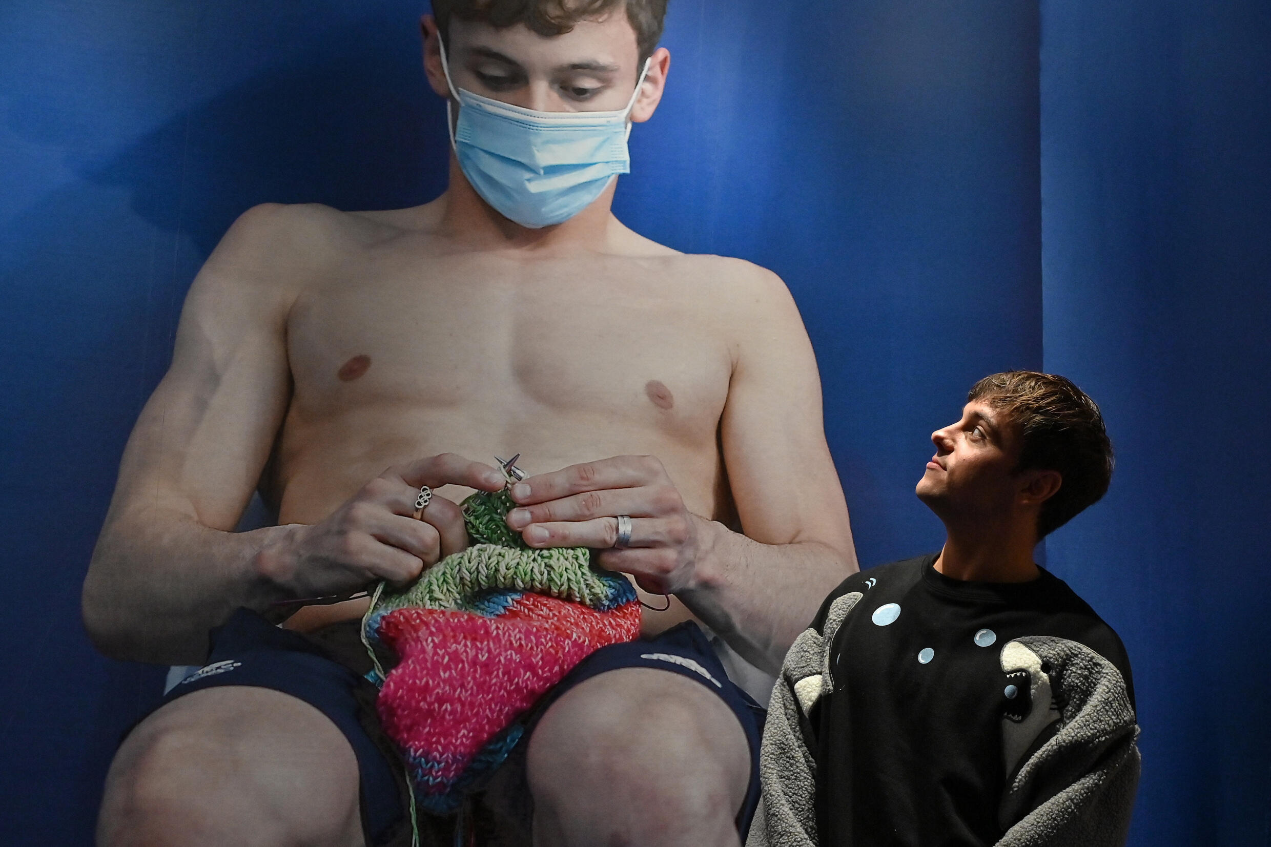 Retired British Olympic diving champion Tom Daley poses next to a photo of himself knitting at a swim meet, at the entrance to a new exhibition showcasing creations he has made since taking up the craft in 2020, after an interview with AFP in the Shibuya district of central Tokyo on November 8, 2024. Photo: AFP